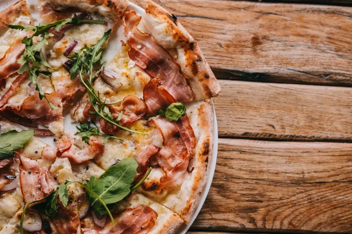 Pyszna pizza carbonara z chrupiącym boczkiem i rukolą, podana na drewnianym blacie.