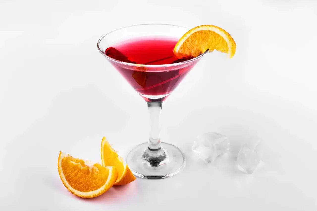 Elegancki, czerwony drink cosmopolitan z plasterkiem pomarańczy, obok kostki lodu i pomarańczy.