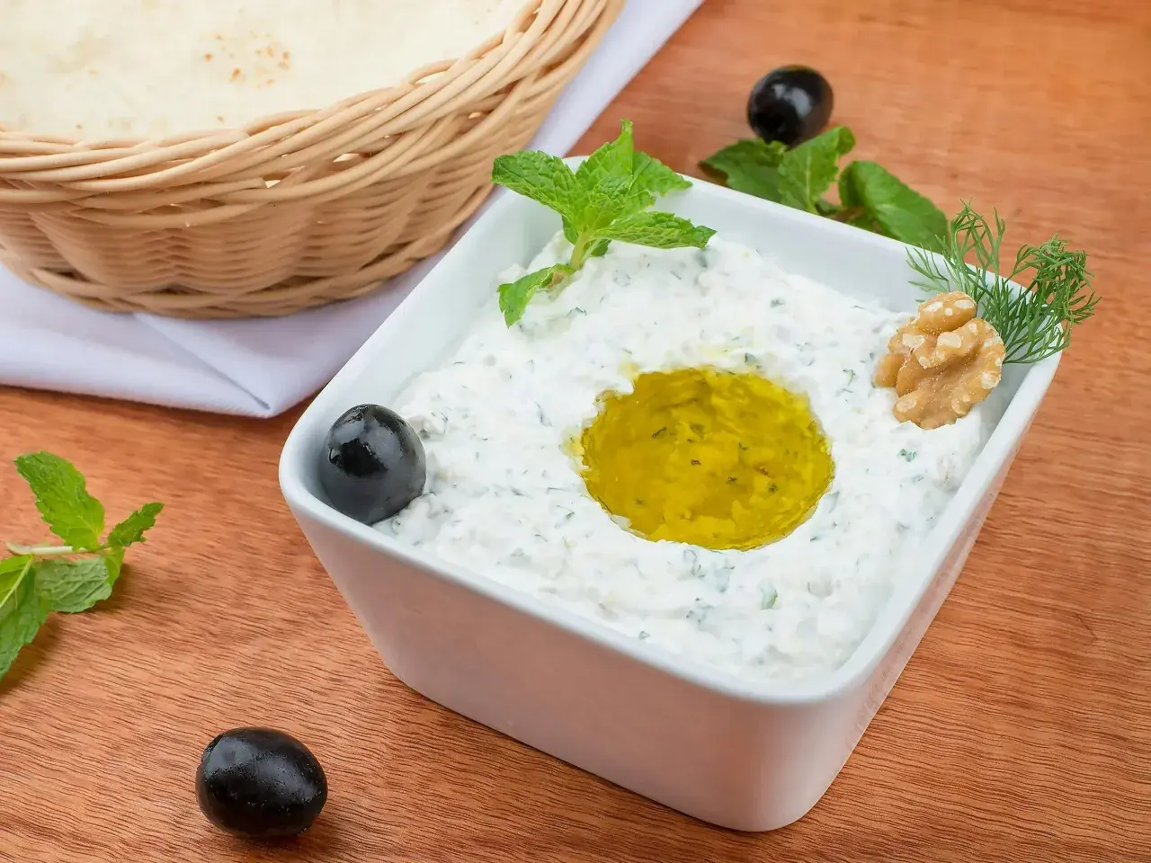 Jak zrobić tzatziki? Kremowy dip z jogurtu, czosnku i ziół, polany oliwą, z oliwkami i orzechami. Obok pieczywo.