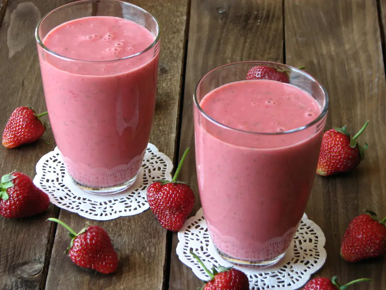 Dwa szklanki orzeźwiającego koktajlu z mrożonych truskawek, otoczone świeżymi owocami na drewnianym tle.