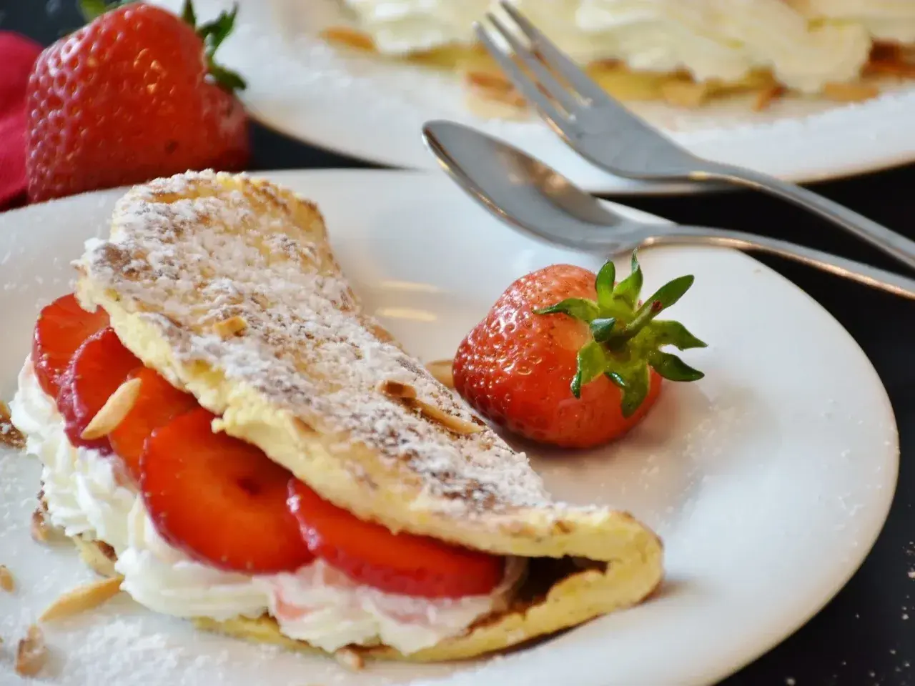 Puszysty omlet z bitą śmietaną i truskawkami, posypany cukrem pudrem i płatkami migdałów. Obok leży świeża truskawka.