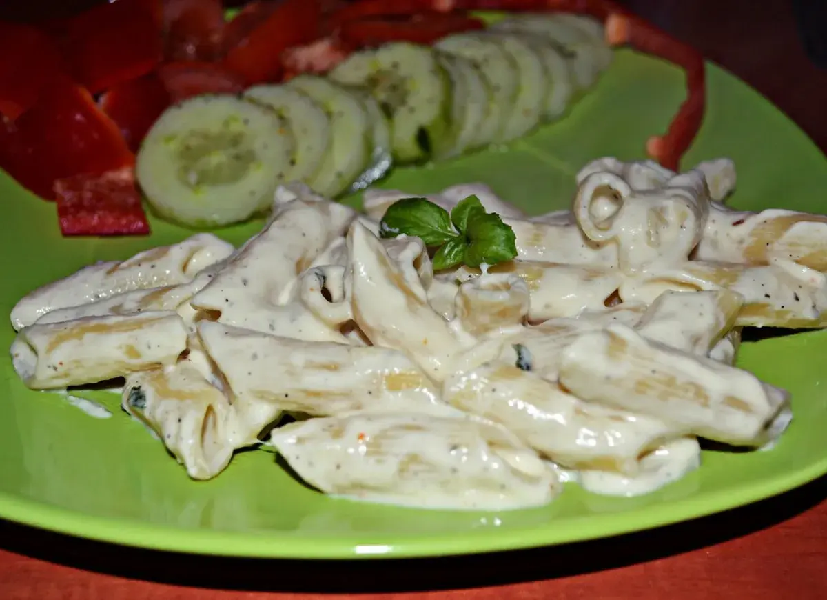Penne w kremowym sosie z mozzarellą, udekorowane bazylią. Obok plasterki ogórka i papryka.