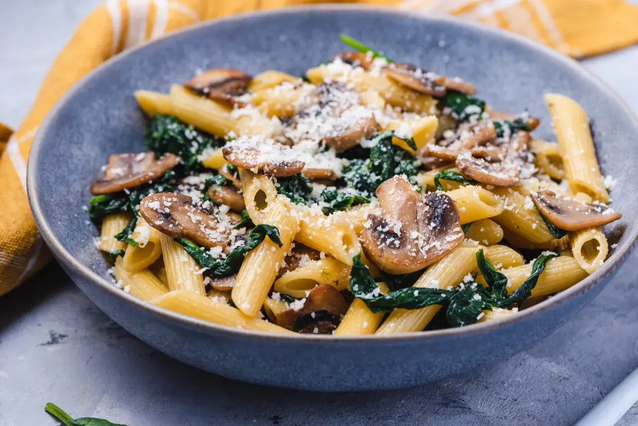 Penne z pieczarkami i szpinakiem, posypane parmezanem, w szarej misce.