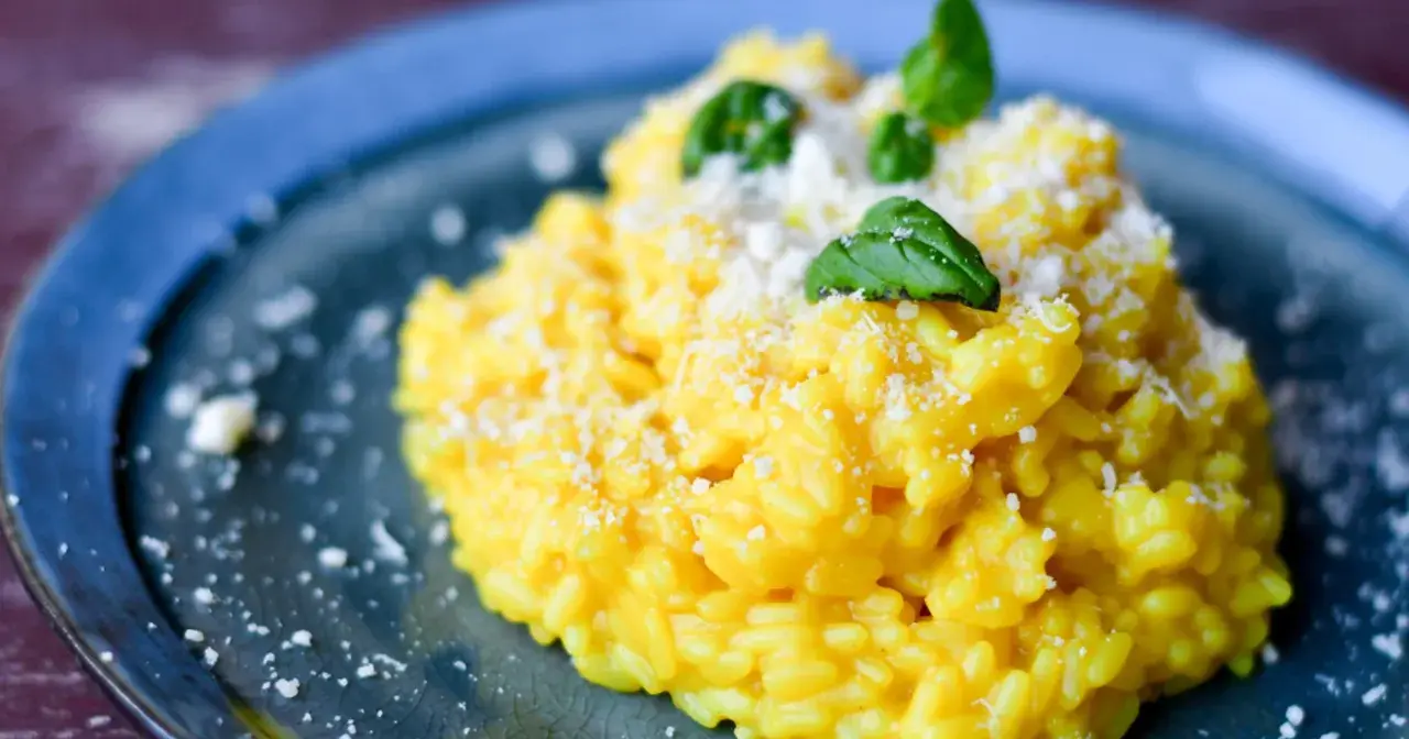 Aksamitny, żółty ryż do risotto, posypany parmezanem i udekorowany listkami bazylii, na niebieskim talerzu.