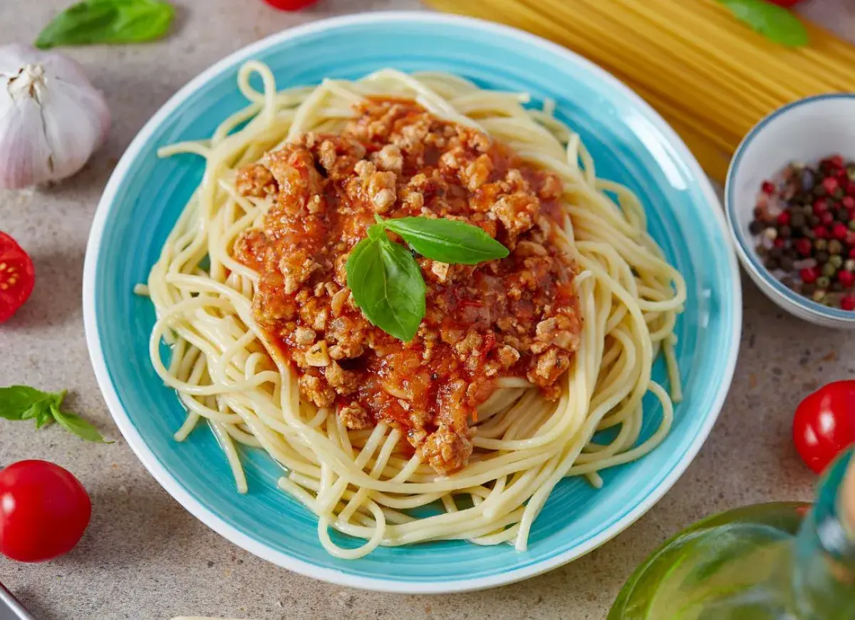 Pyszne spaghetti bolognese z passaty, podane na turkusowym talerzu, ozdobione listkiem bazylii. Obok czosnek, pomidorki i kolorowy pieprz.