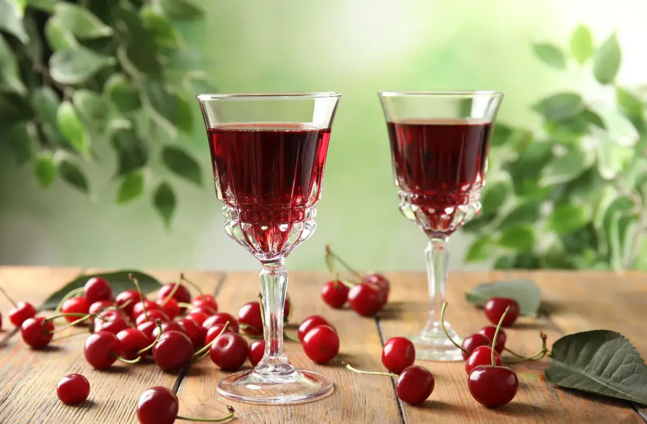 Dwa kieliszki z rubinową, szybką nalewką z wiśni na spirytusie, otoczone świeżymi wiśniami na drewnianym stole.