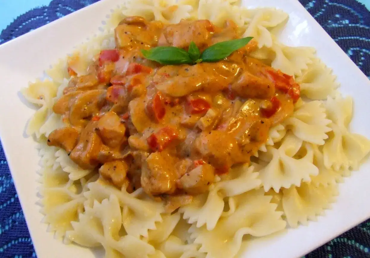 Pyszny kurczak w sosie paprykowym z pomidorami, podany na makaronie farfalle. Udekorowany listkiem bazylii.