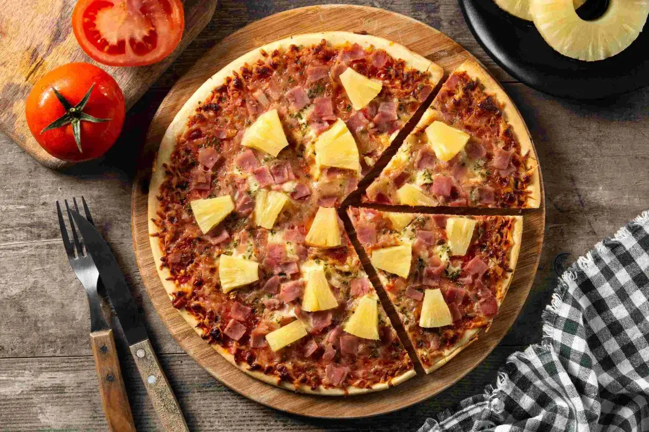 Pyszna pizza z ananasem i szynką, podana na drewnianej desce. Obok leżą pomidory i sztućce.