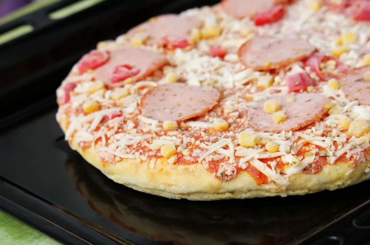Gotowa do pieczenia pizza mrożona na czarnej blasze. Widoczne plastry salami, kukurydza i ser.