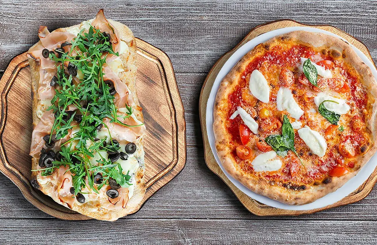 Pinsa co to? Dwa przysmaki: prostokątna pinsa z szynką, rukolą i oliwkami oraz okrągła pizza Margherita z pomidorkami i mozzarellą.