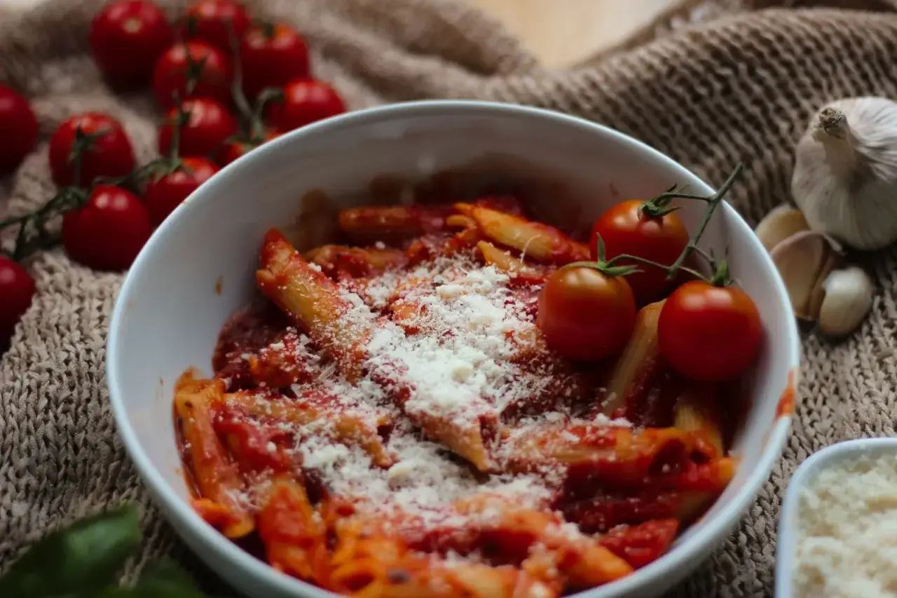 Penne z aromatycznym sosem pomidorowym do makaronu, posypane parmezanem. Obok świeże pomidorki i czosnek.