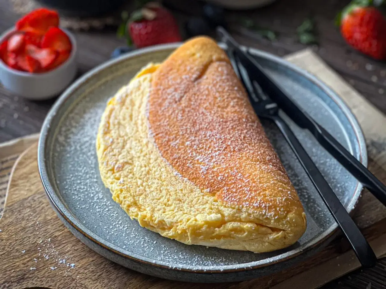 Puszysty omlet, lekko przyprószony cukrem pudrem, czeka na talerzu. Obok czarne sztućce i miseczka z truskawkami.