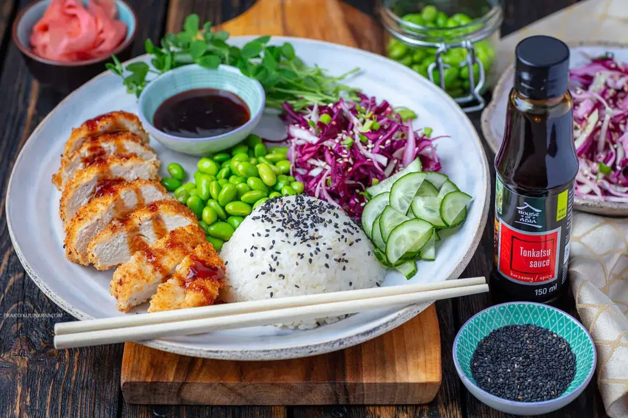 Pyszny ryż z kurczakiem w panierce, podany z edamame, czerwoną kapustą, ogórkiem i sosem tonkatsu.