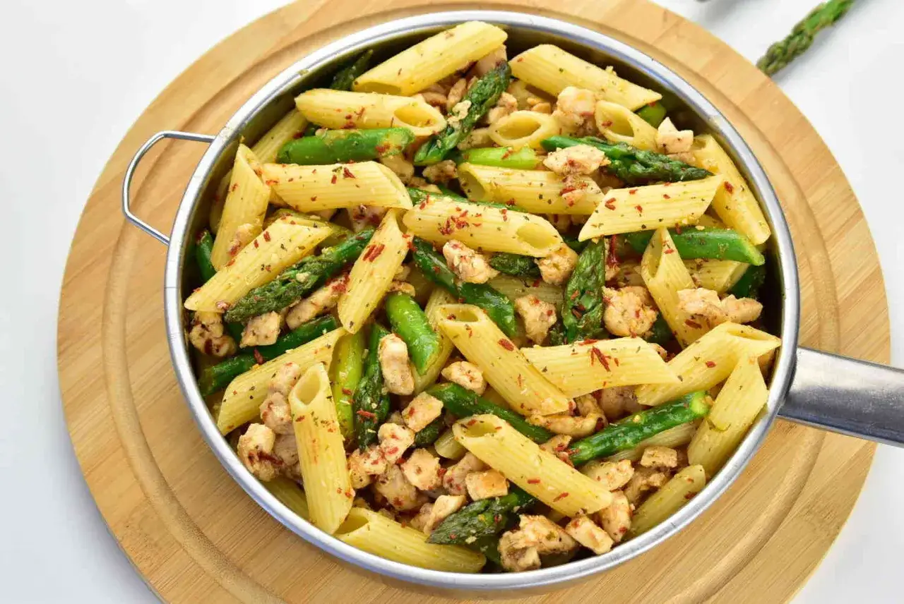 Penne z kurczakiem i szparagami, posypane płatkami chili, w metalowej misce na drewnianej desce.