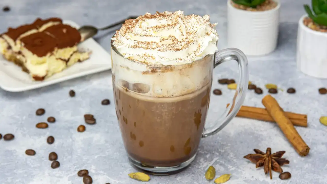 Aromatyczna kawa z kakao, udekorowana bitą śmietaną i posypką, obok tiramisu i przypraw.