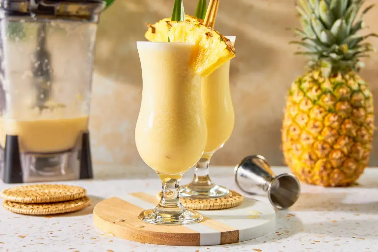 Dwa orzeźwiające koktajle Pina Colada, udekorowane ananasem, obok blendera i świeżego owocu. Idealny przepis na lato!