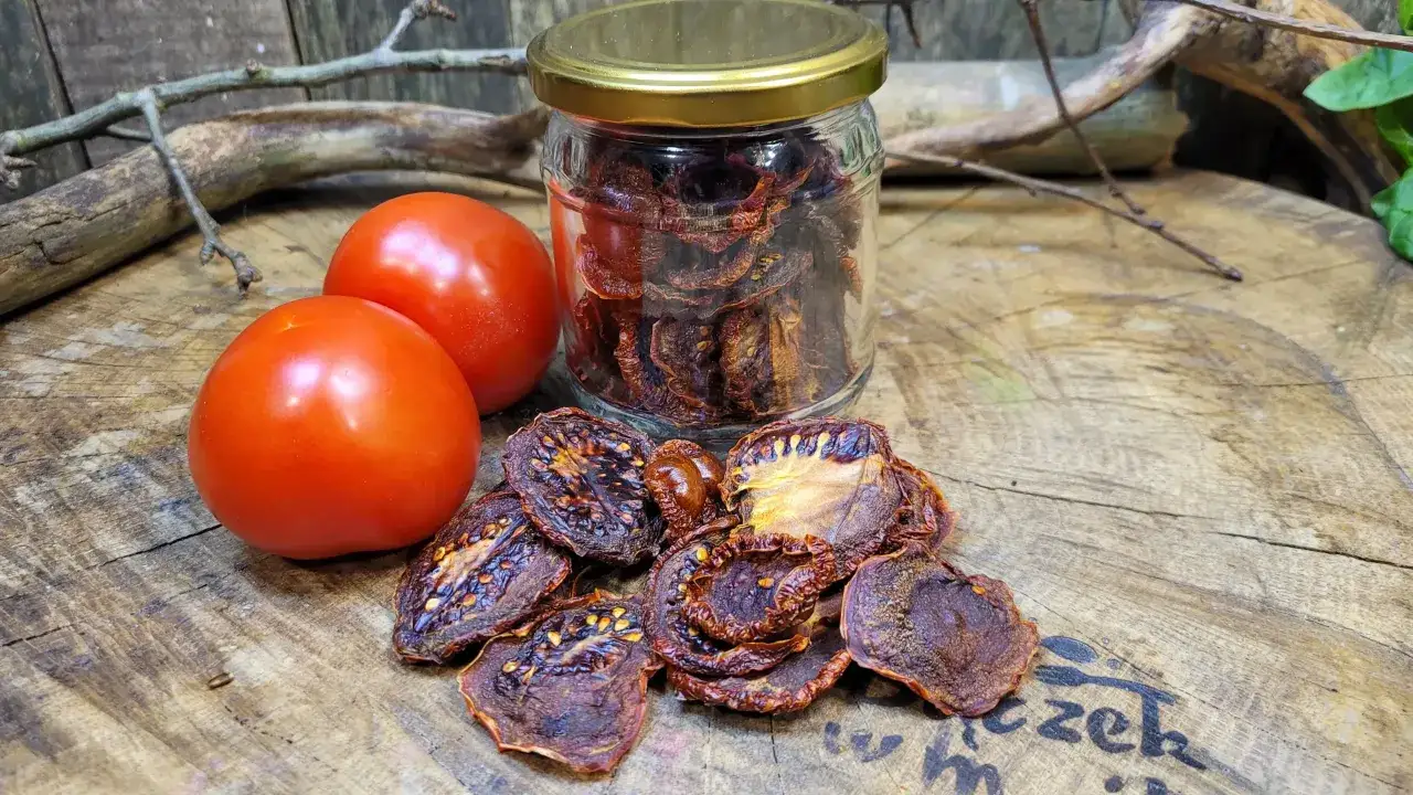 Słoik pełen aromatycznych suszonych pomidorów obok świeżych, czerwonych pomidorów i gałązek na drewnianym blacie.