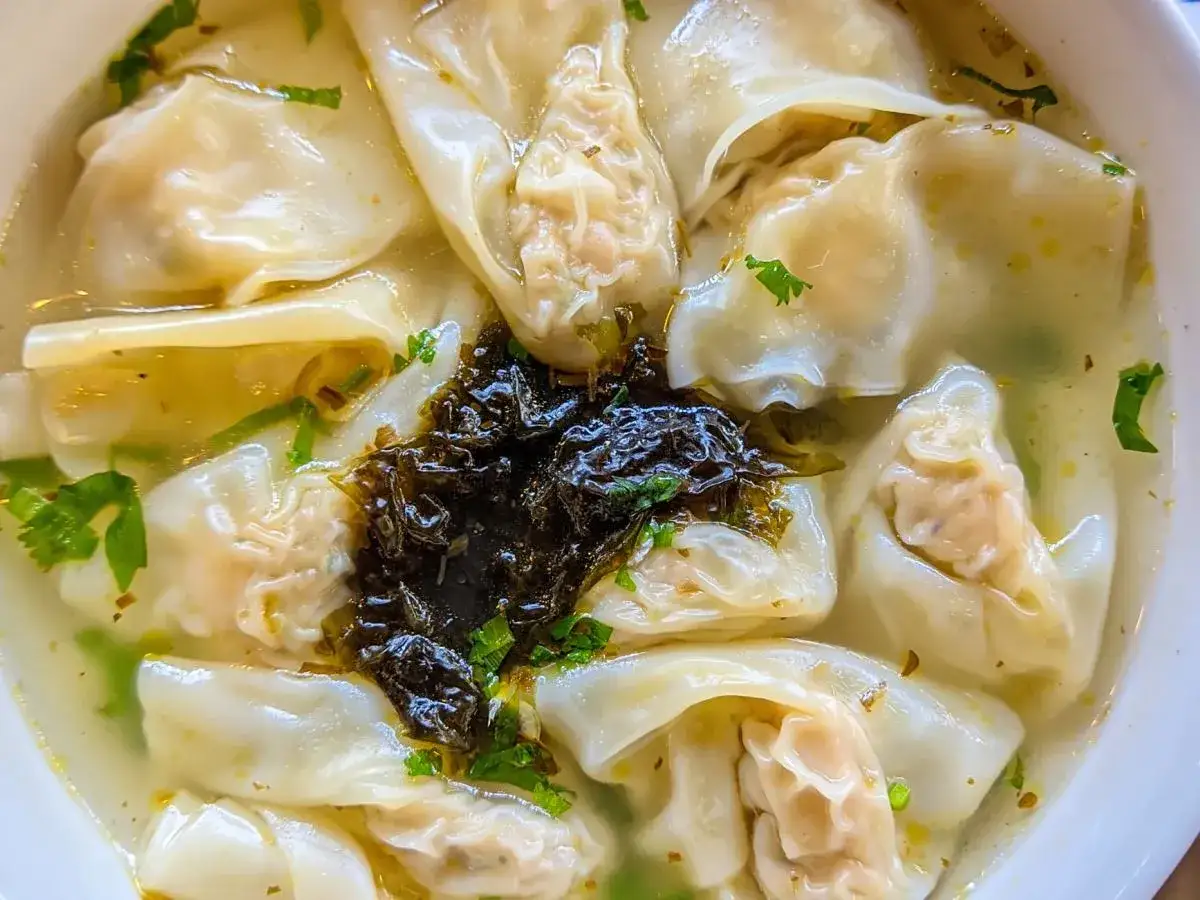 A miseczka pełna aromatycznej zupy won ton, z delikatnymi pierożkami pływającymi w bulionie, ozdobiona zieloną natką i ciemną pastą.