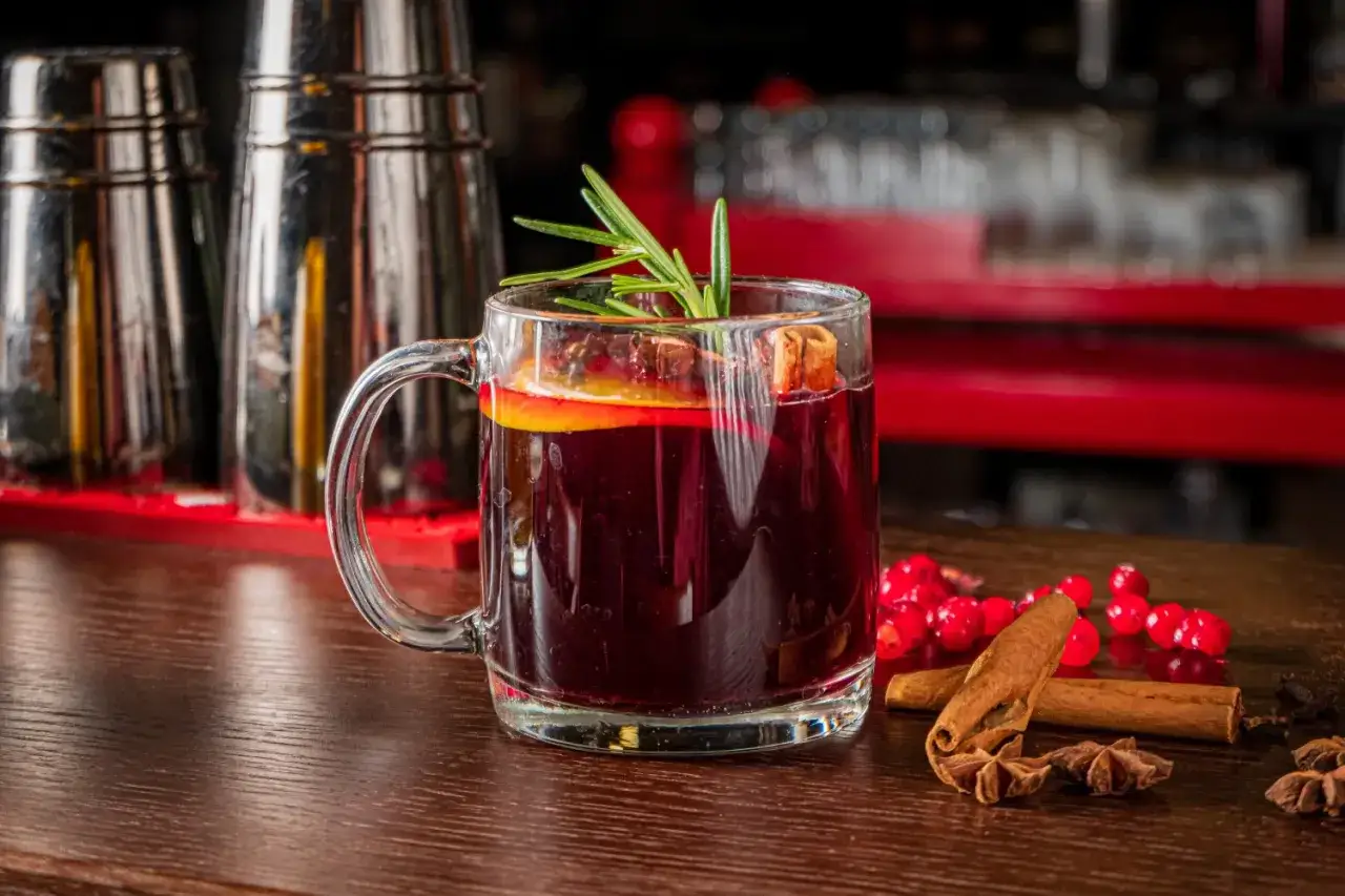 Aromatyczny grog w szklanej kuflu, ozdobiony pomarańczą i rozmarynem, obok cynamonu i żurawiny.
